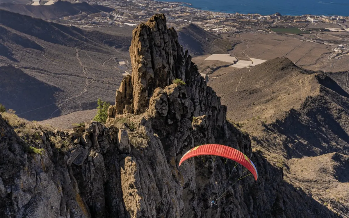 Uma asa de parapente vermelha a passar por um pico rochoso acima de vales em socalcos no sul de Tenerife