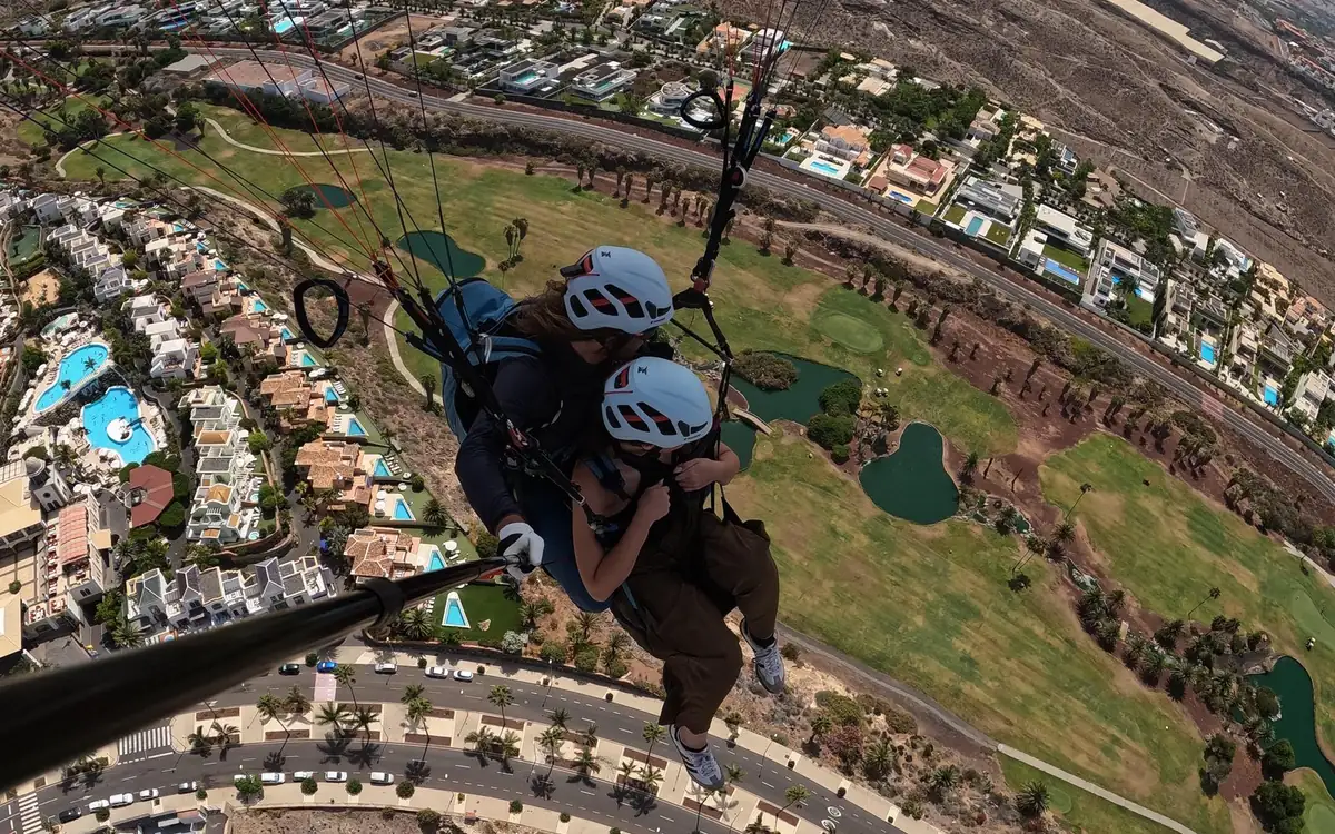 A vista diretamente para baixo entre os pés de dois passageiros de parapente tandem sobre piscinas, campos de golfe e a estrada costeira