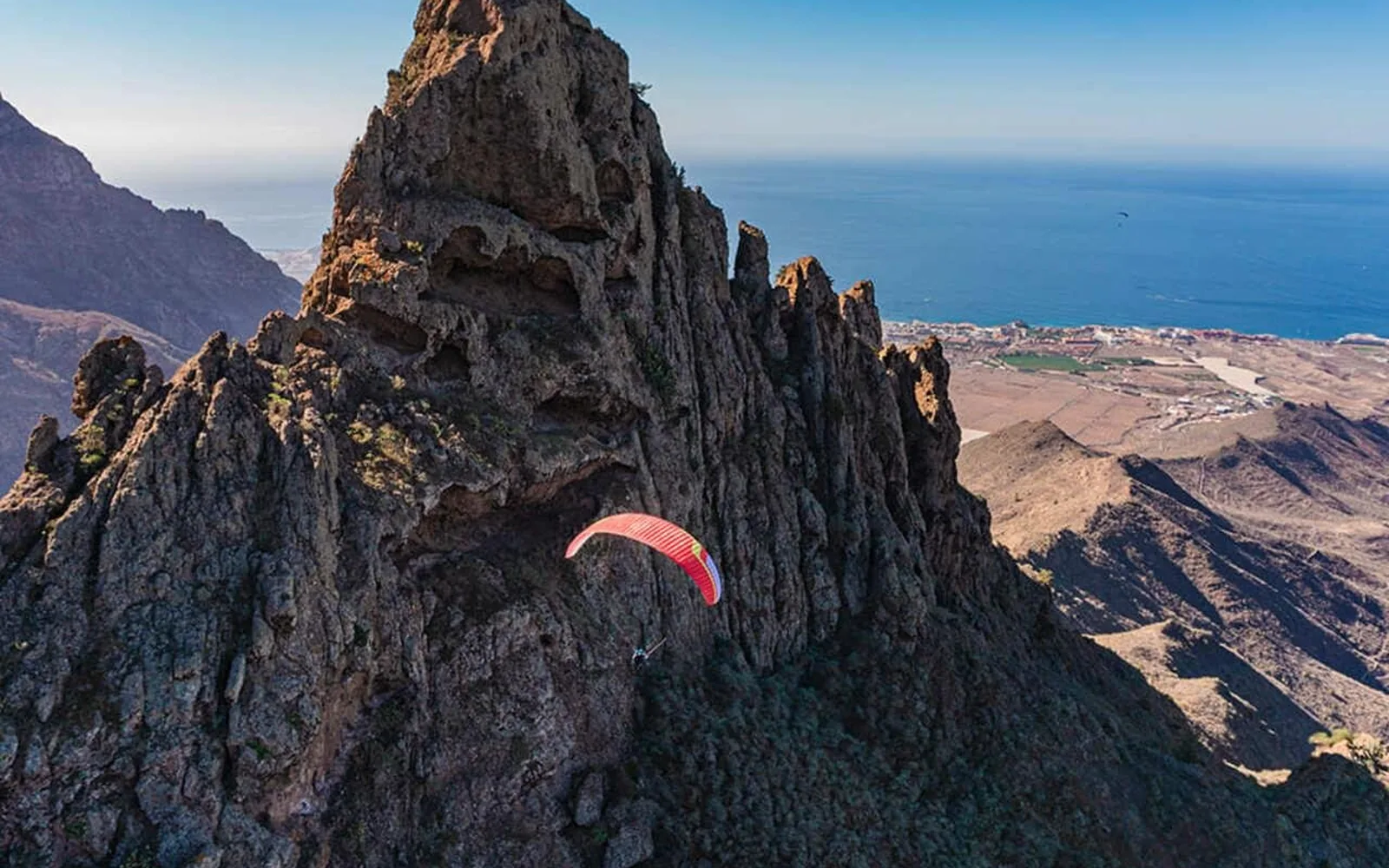 Uma asa de parapente vermelha ao lado de uma crista vulcânica recortada, com a planície costeira e o Atlântico muito abaixo