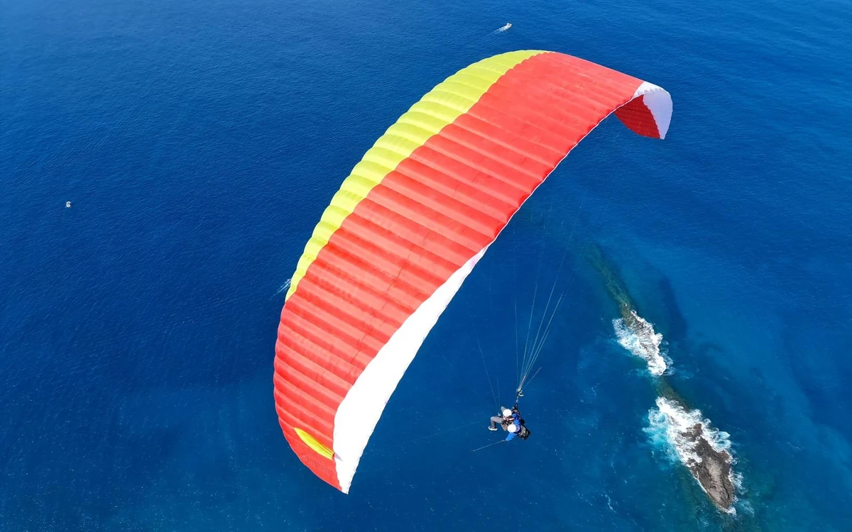 Um parapente visto de cima sobre água aberta azul-escura com rebentação nas rochas em baixo