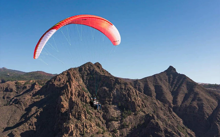 Uma asa vermelha a transportar um par tandem sobre picos rochosos nus sob um céu limpo