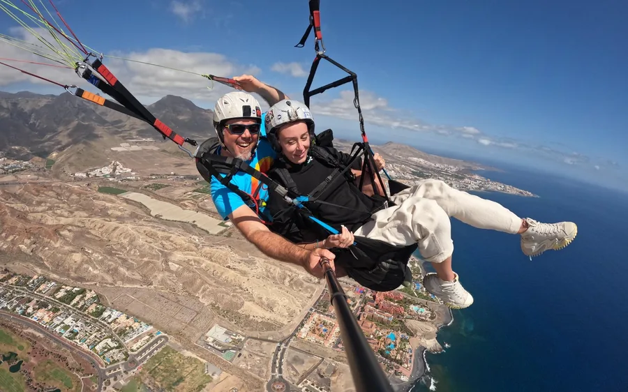 Um piloto e um passageiro a sorrir num pau de selfie bem alto sobre a planície costeira seca e os resorts