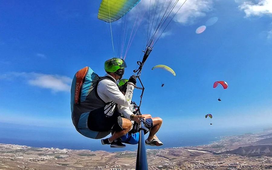 Vistos de trás, um piloto e um passageiro com capacetes verdes sob uma asa amarelo-esverdeada, mais três asas no azul e a costa sul abaixo